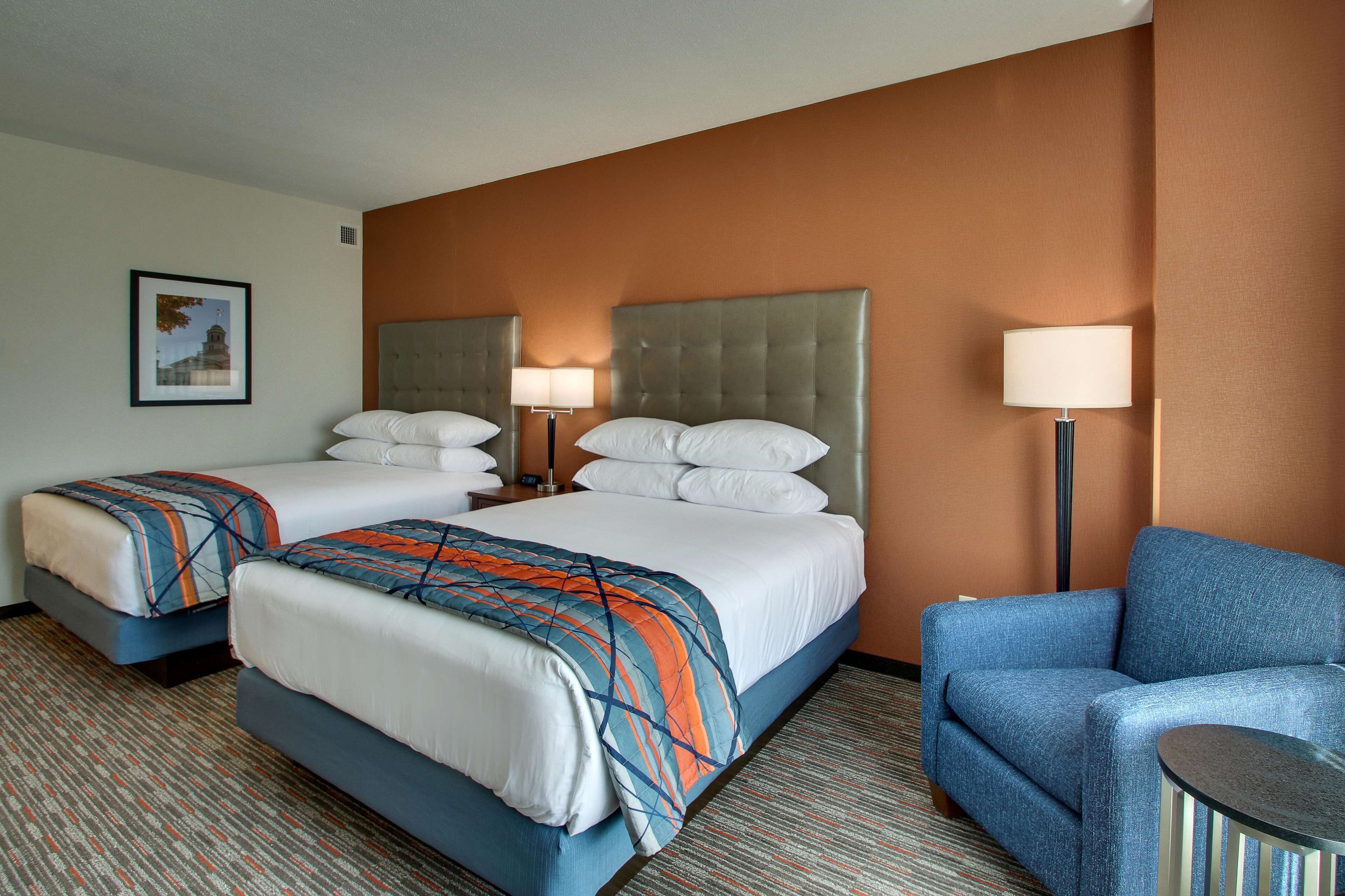 A hotel room with two beds, a blue armchair, and a small table.