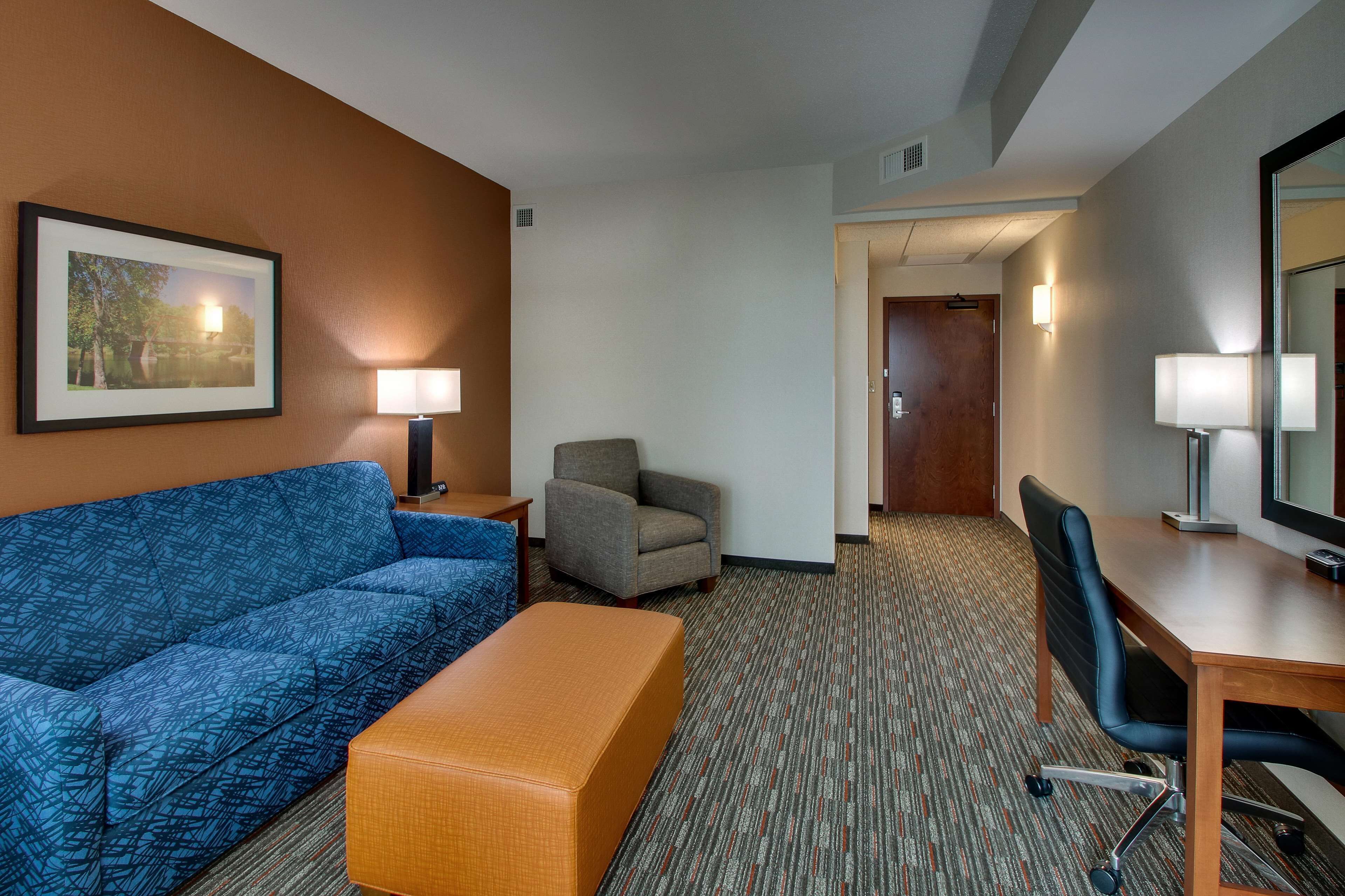 Habitación de hotel con sofá azul, otomana naranja, sillón gris, escritorio de madera y un espejo grande.