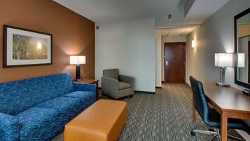 Quarto de hotel com um sofá azul, um banquinho laranja, uma poltrona cinzenta, uma escrivaninha de madeira e um espelho grande.
