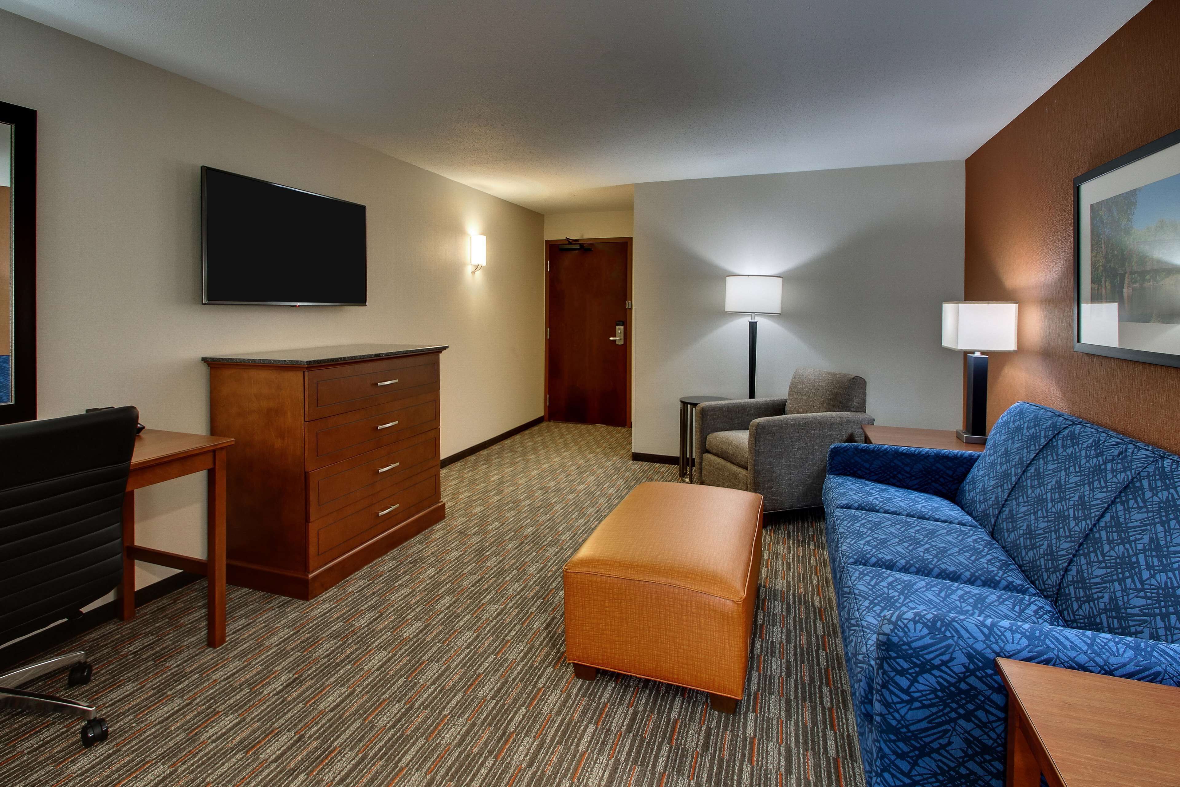 A hotel room with a flat-screen TV, a wooden dresser, a blue sofa, a small table, and a lamp.