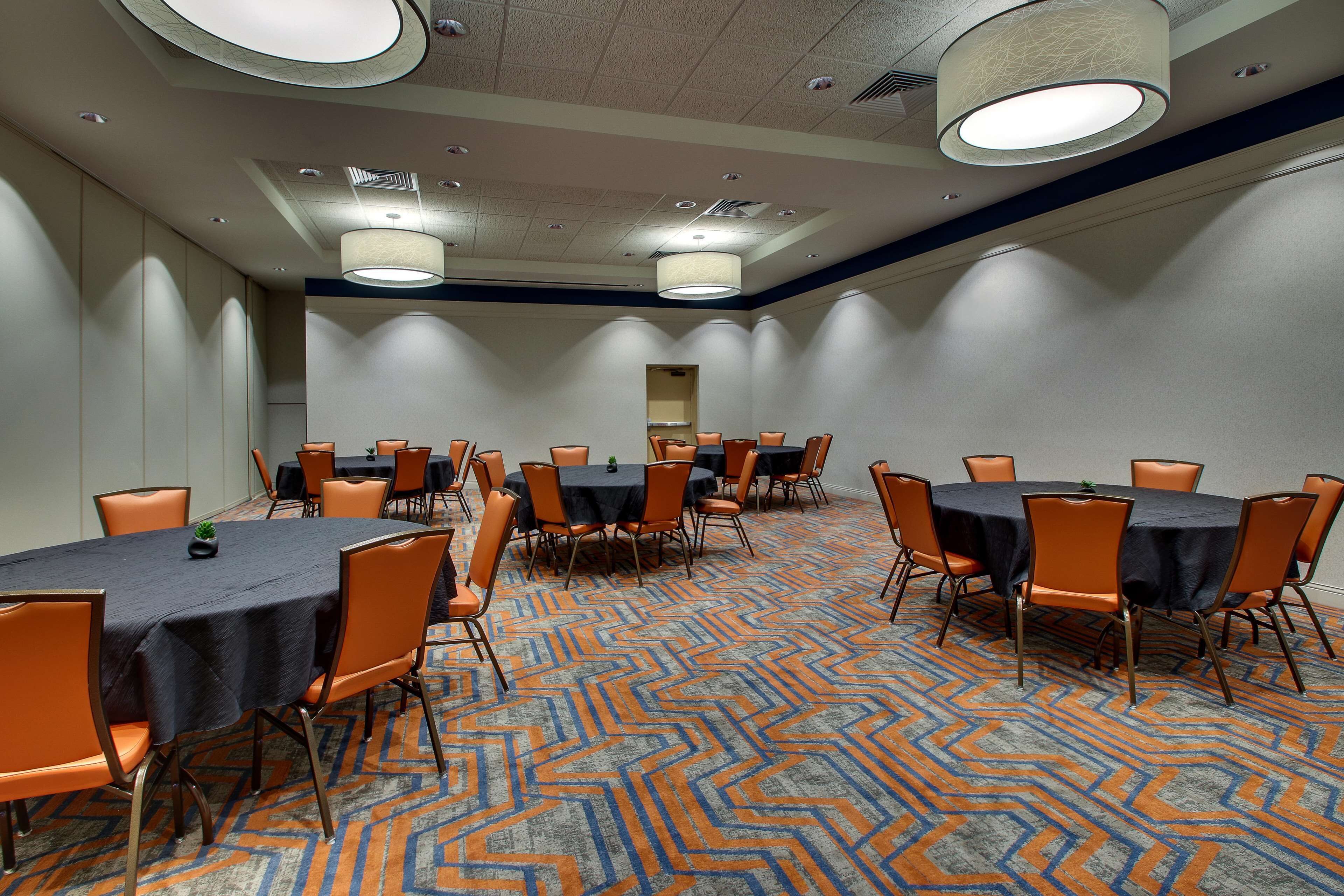 Un salón de conferencias con varias mesas y sillas, suelo alfombrado e iluminación en el techo.