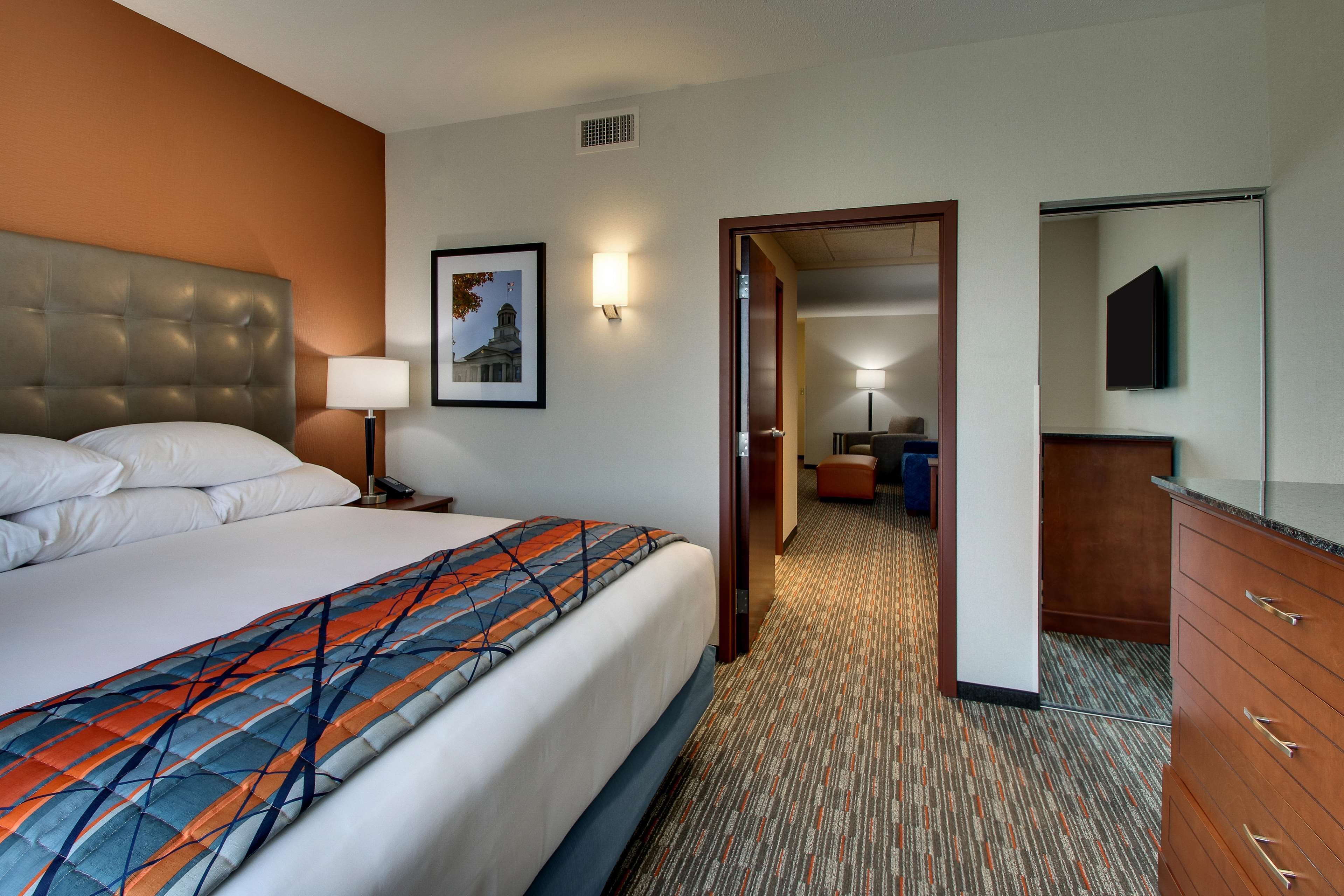 A hotel room with a large bed, a nightstand, a lamp, a framed picture on the wall, a wooden dresser, and a door leading to another room.