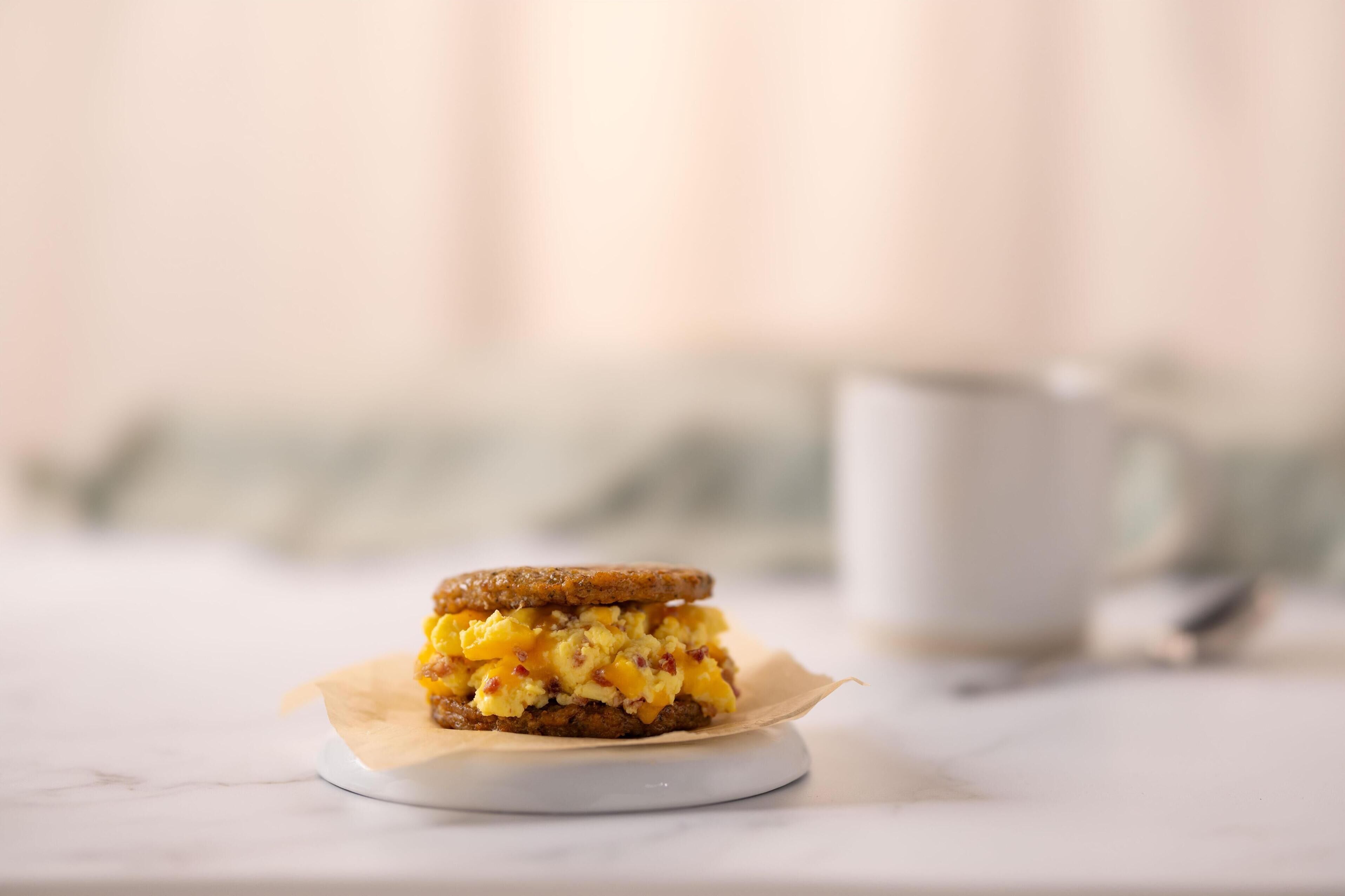 Un sándwich en un plato blanco con una servilleta, una taza blanca y una cuchara al fondo.