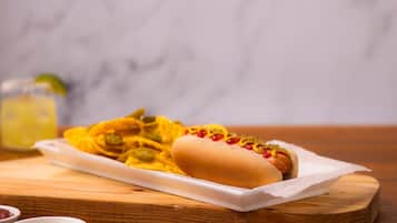 A hot dog with mustard, ketchup, and relish on a bun, accompanied by a side of potato chips and a drink in the background.