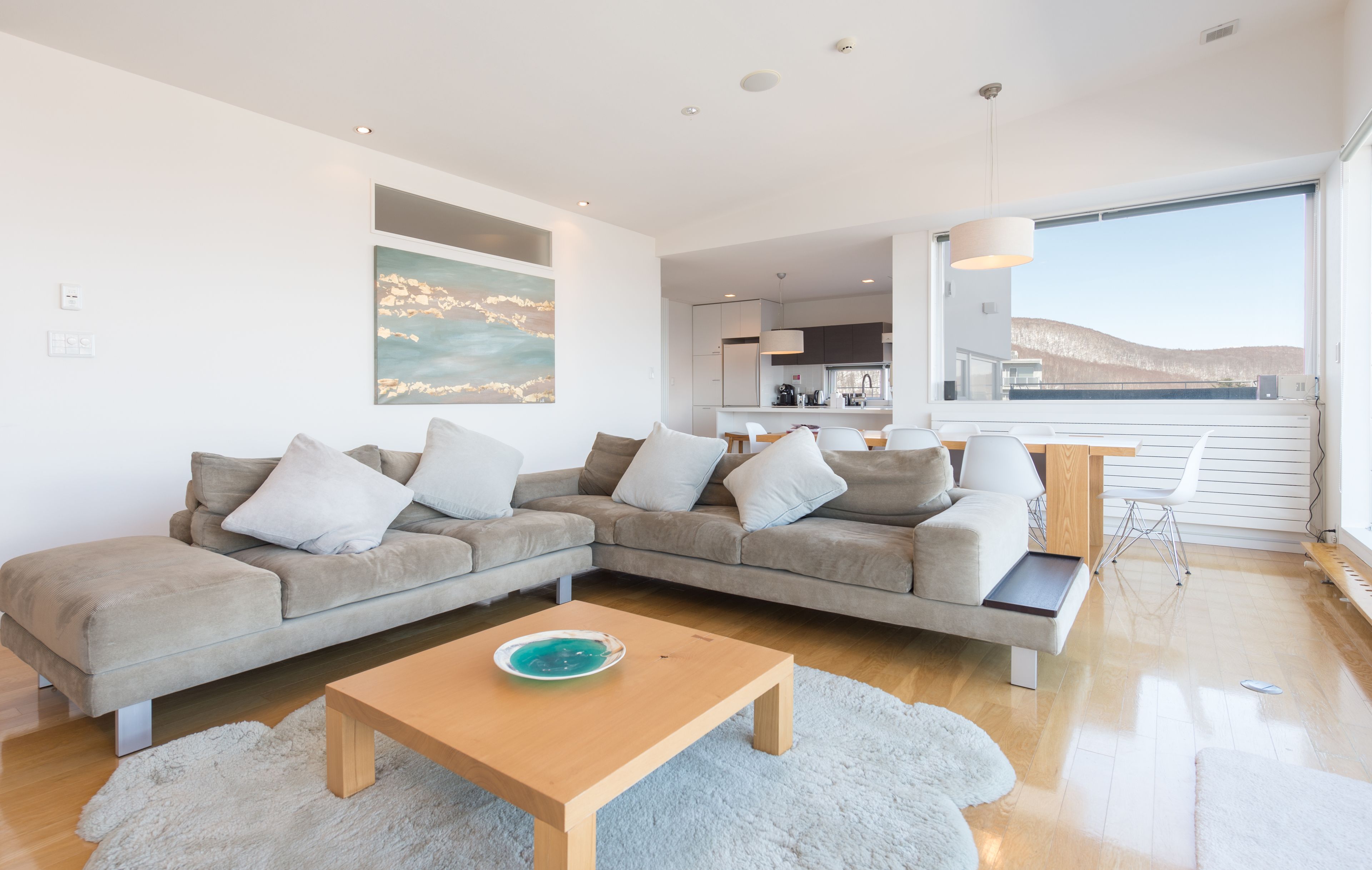 A modern living room with a sectional sofa, a wooden coffee table, and a view of the kitchen and balcony.