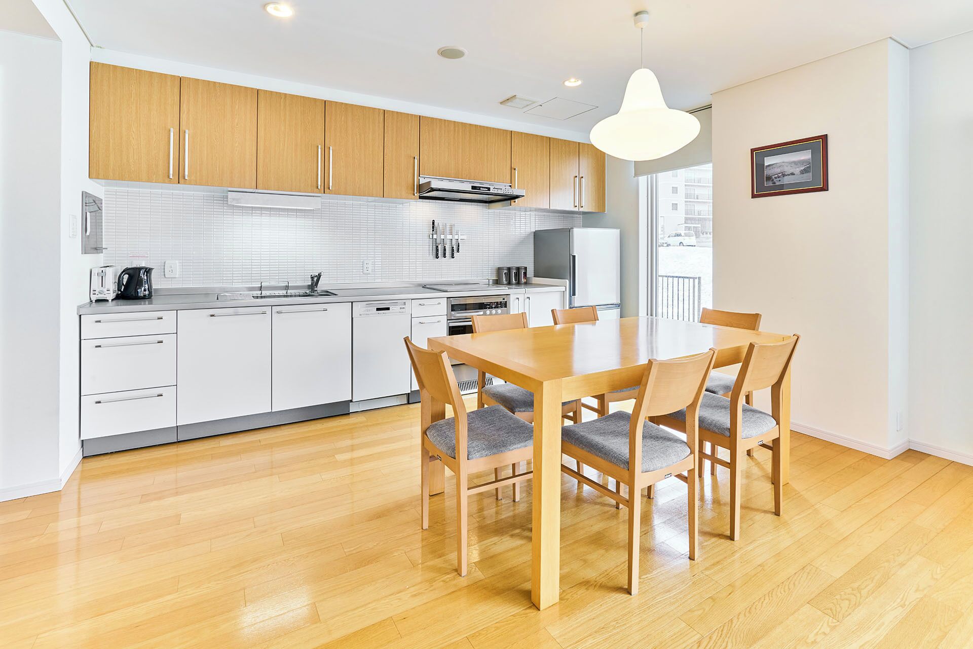 A modern kitchen with white cabinets, a wooden dining table, and a large window.