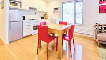 Una cocina moderna con armarios blancos, un microondas empotrado y una estufa. Hay una mesa de comedor con cuatro sillas rojas. Una ventana proporciona luz natural, y un sofá es visible al fondo.
