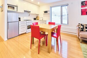 Una cocina moderna con armarios blancos, un microondas empotrado y una estufa. Hay una mesa de comedor con cuatro sillas rojas. Una ventana proporciona luz natural, y un sofá es visible al fondo.