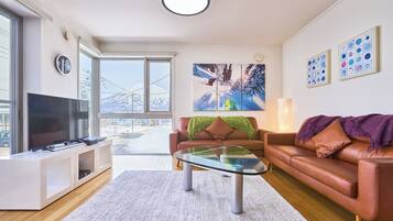 A modern living room with a glass coffee table, leather sofas, a flat-screen TV, and large windows offering a view of a snowy landscape.