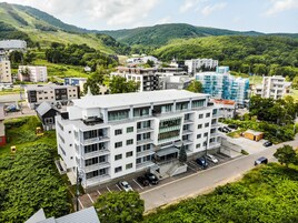 Un bâtiment moderne à plusieurs étages, doté d’un toit plat, entouré d’une végétation luxuriante et avec des collines en arrière-plan.