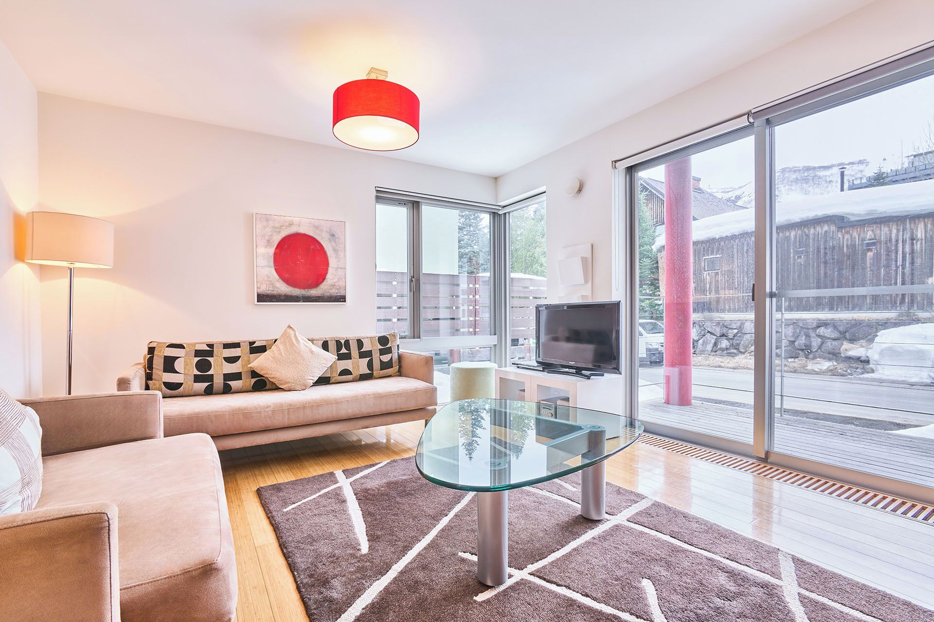 A modern living room with a sofa, a glass coffee table, a TV, and large windows offering a view of the outside.