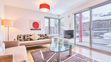 A modern living room with a sofa, a glass coffee table, a TV, and large windows offering a view of the outside.