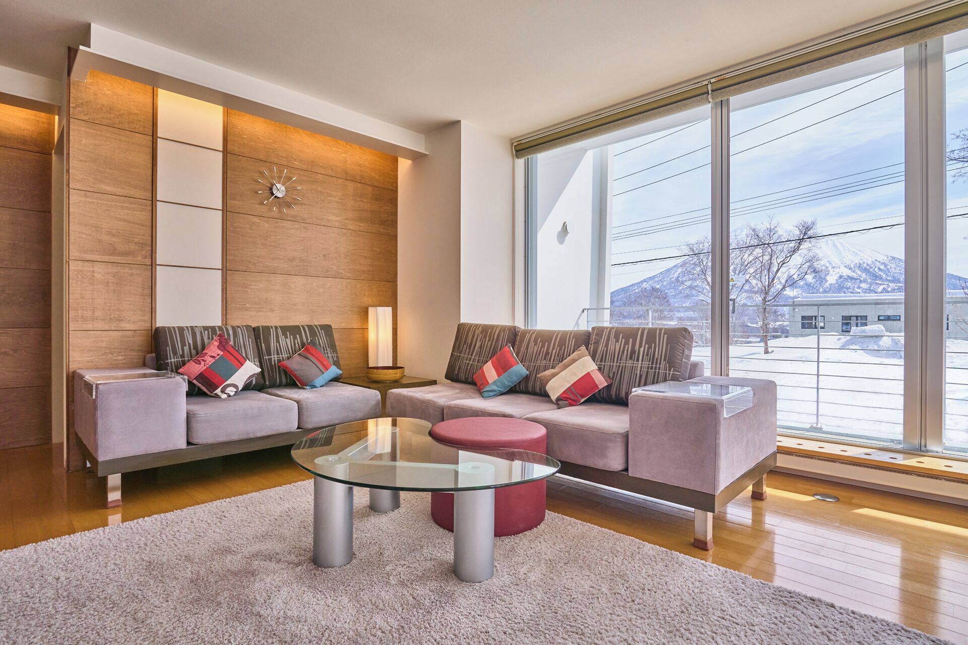 Una sala de estar moderna con una mesa de centro de vidrio, dos sofás con cojines de colores, un ventanal con vista a un paisaje nevado y un reloj fijado a la pared.