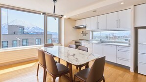 A modern kitchen with a marble table and four chairs, large windows with a mountain view, and white cabinetry. - Niseko Central Condominiums (Kutchan)