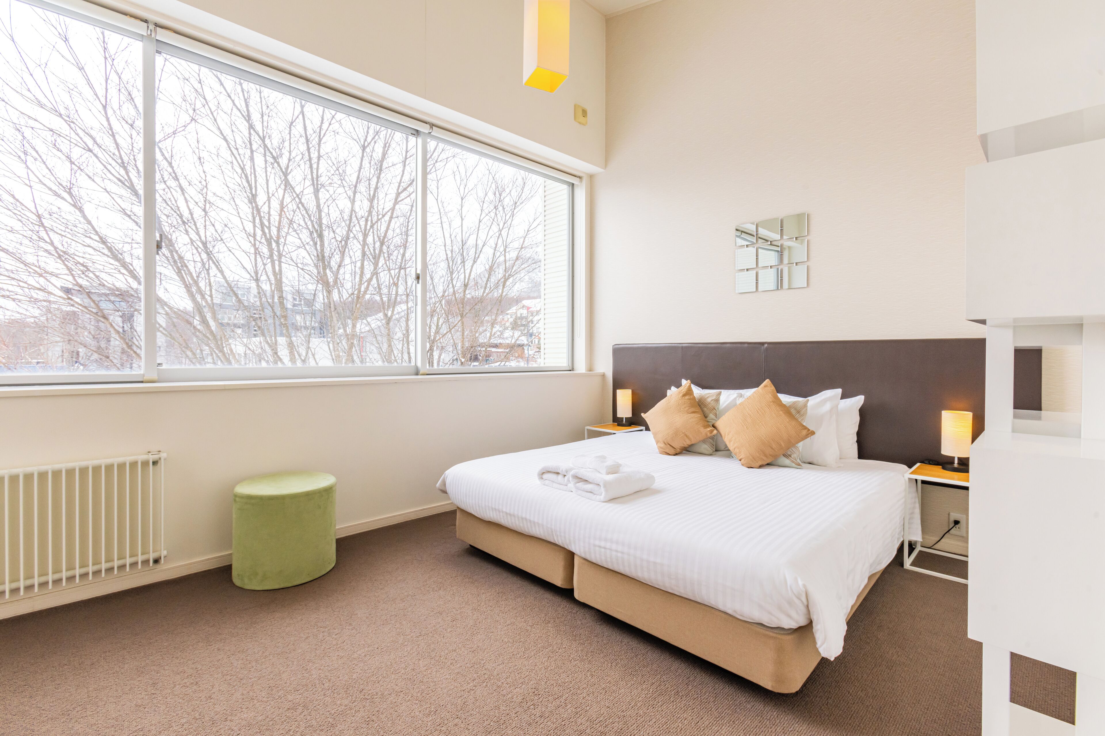 A hotel room with a large window, a bed with white bedding, a green ottoman, and a nightstand with a lamp.