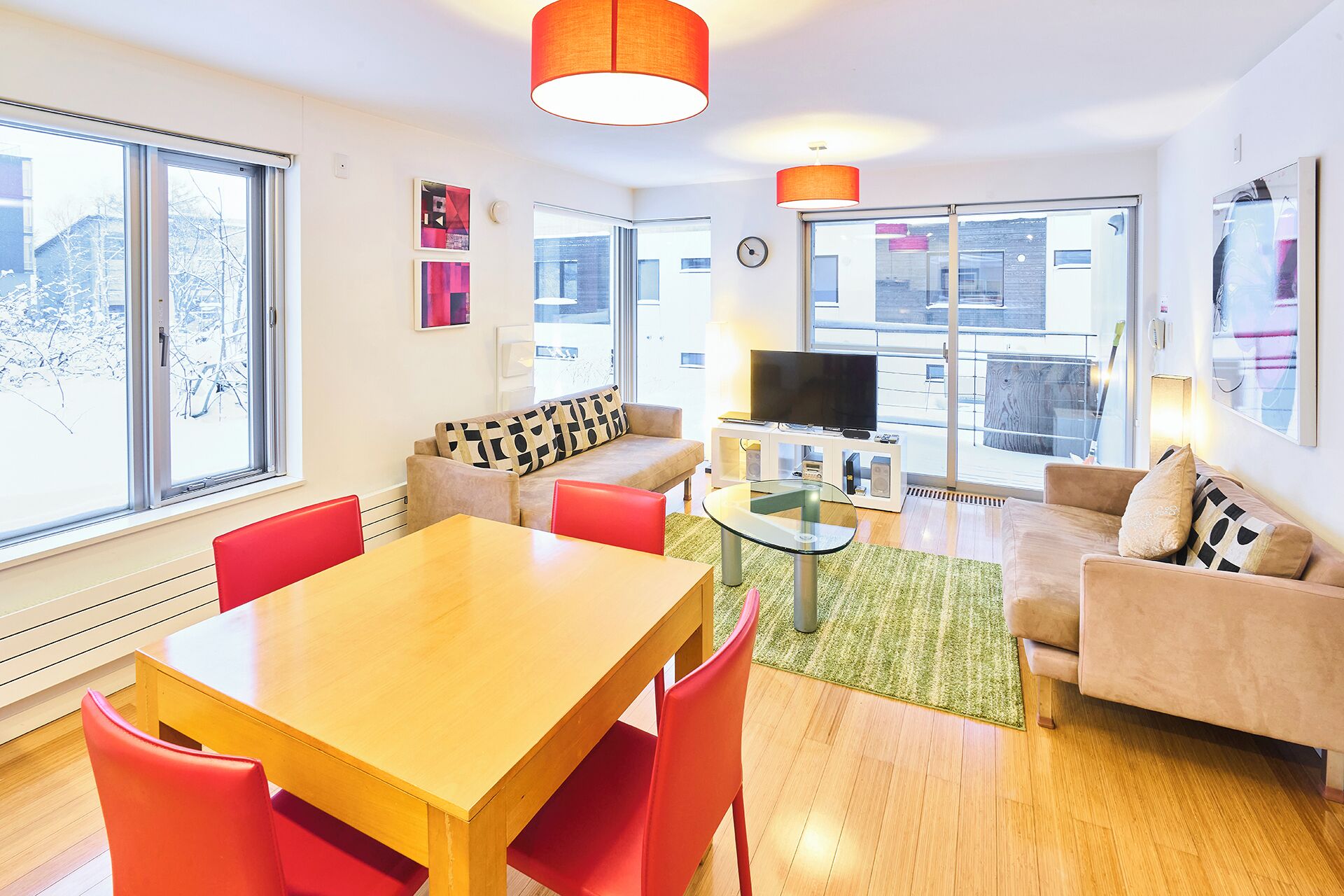 A modern living room with a wooden dining table, red chairs, a beige sofa, and a TV on a white stand.