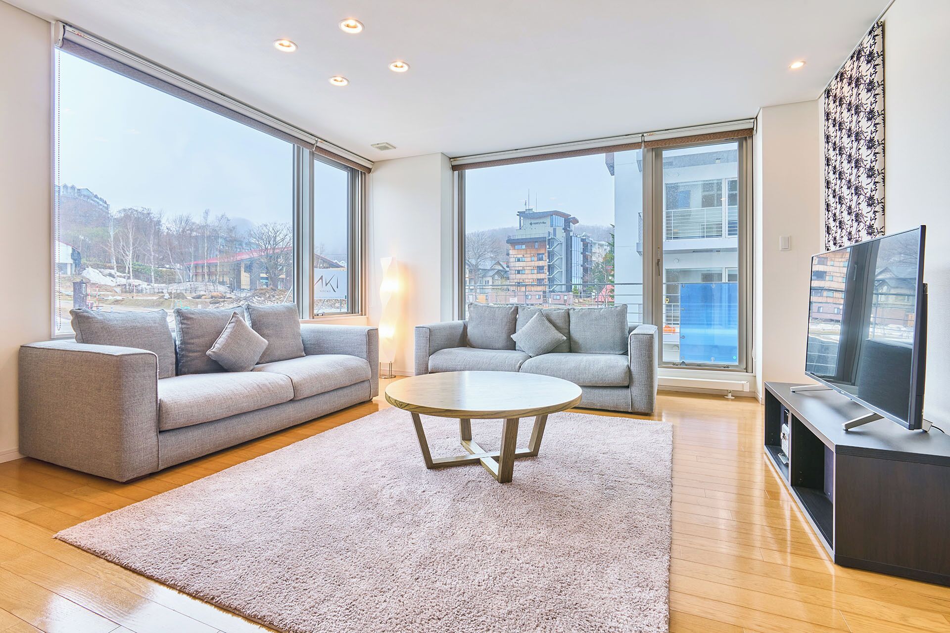 A modern living room with a grey sofa, a round coffee table, and a TV on a stand. Large windows offer a view of buildings and a cityscape.