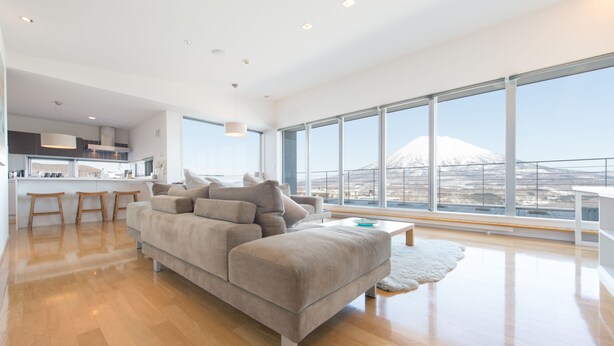 A modern living room with a large gray sofa, wooden flooring, and a view of a mountain through large windows. - Niseko Central Condominiums (Kutchan)