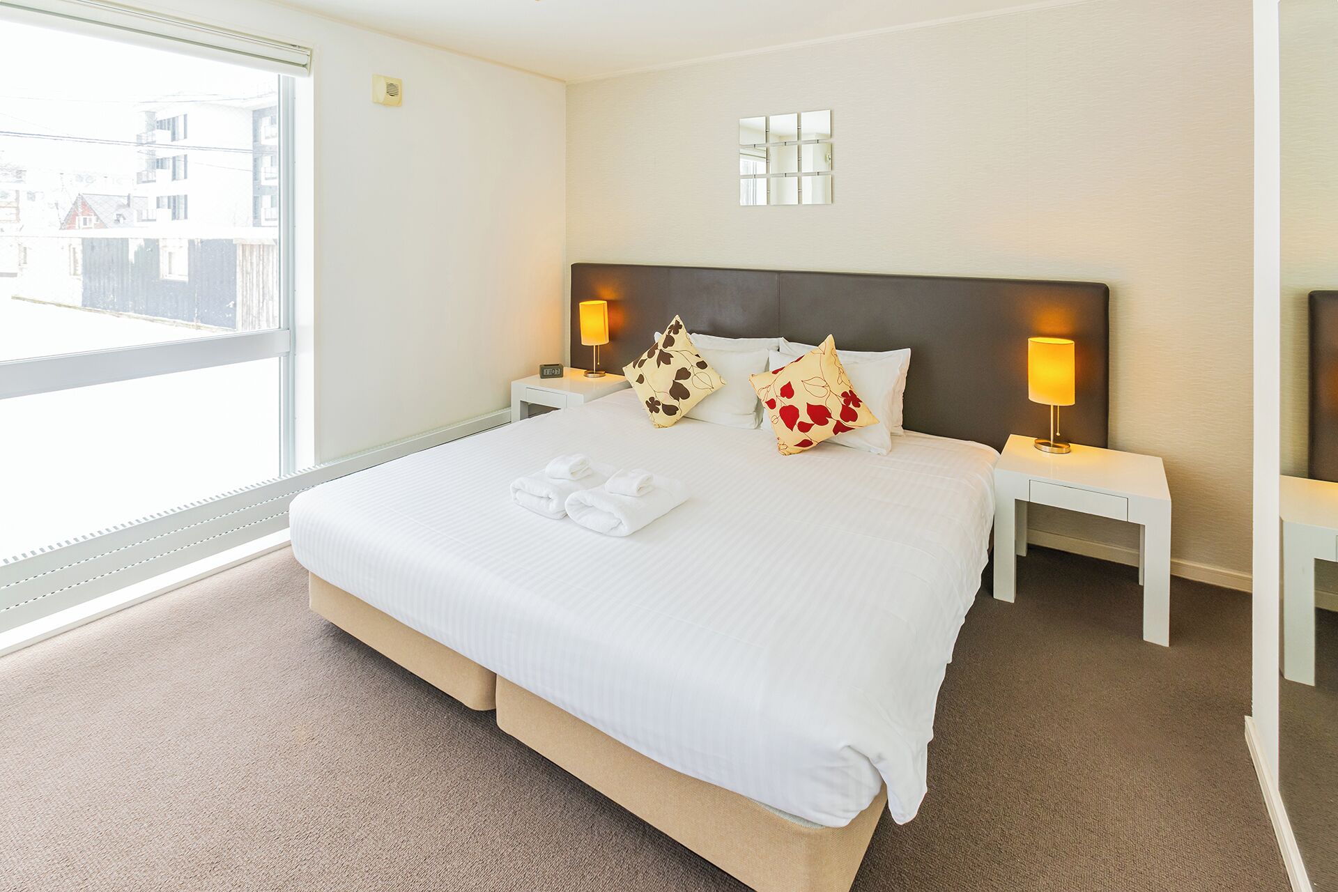 A hotel room with a large bed, two bedside tables, a nightstand, a window with a view, and a wall-mounted mirror.