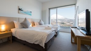 A hotel room with two beds, a desk, a TV, and a large window with a mountain view. - Niseko Central Condominiums (Kutchan)