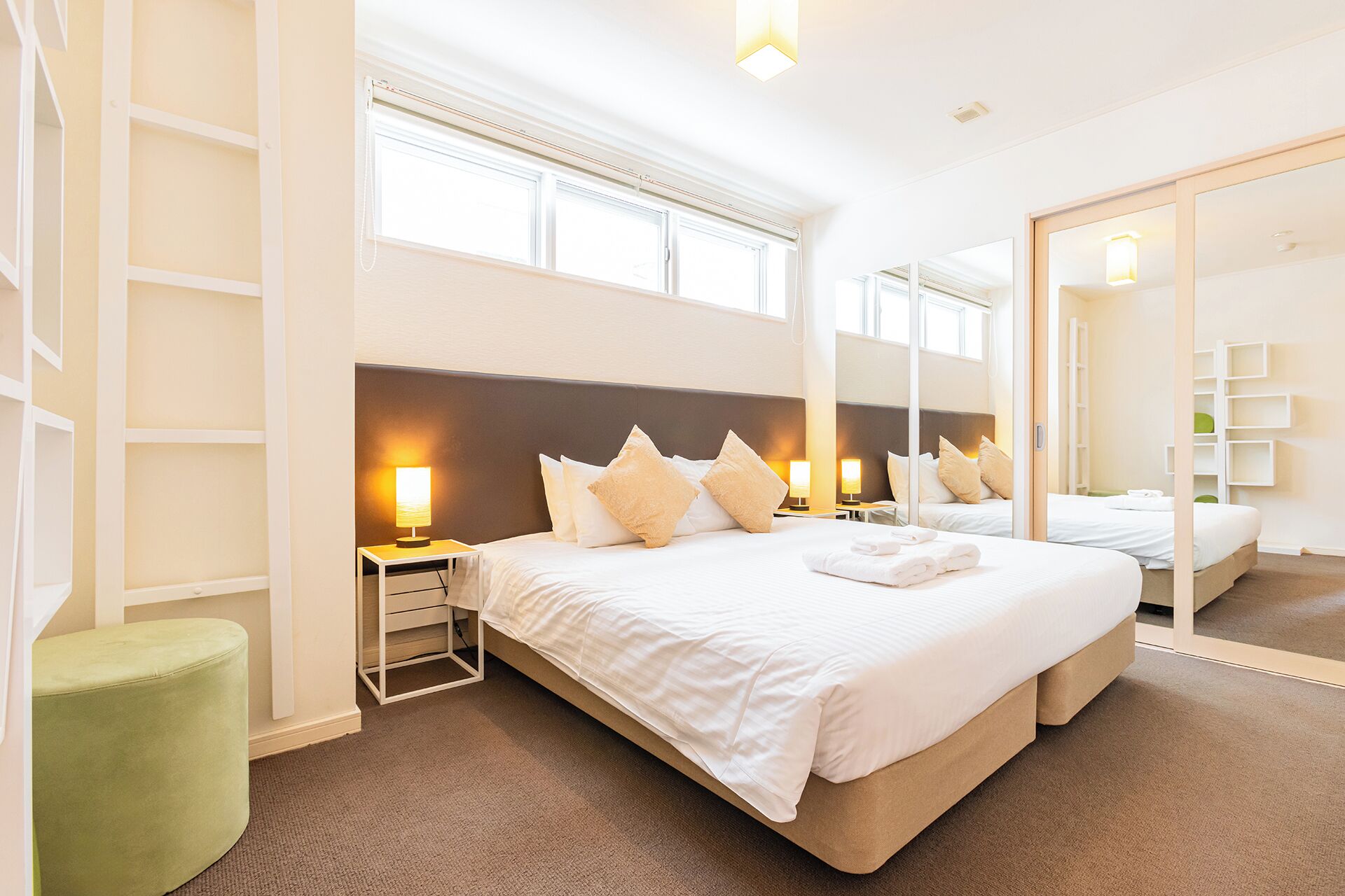 A hotel room with a large bed, bedside lamps, a green ottoman, and a mirror reflecting the room.