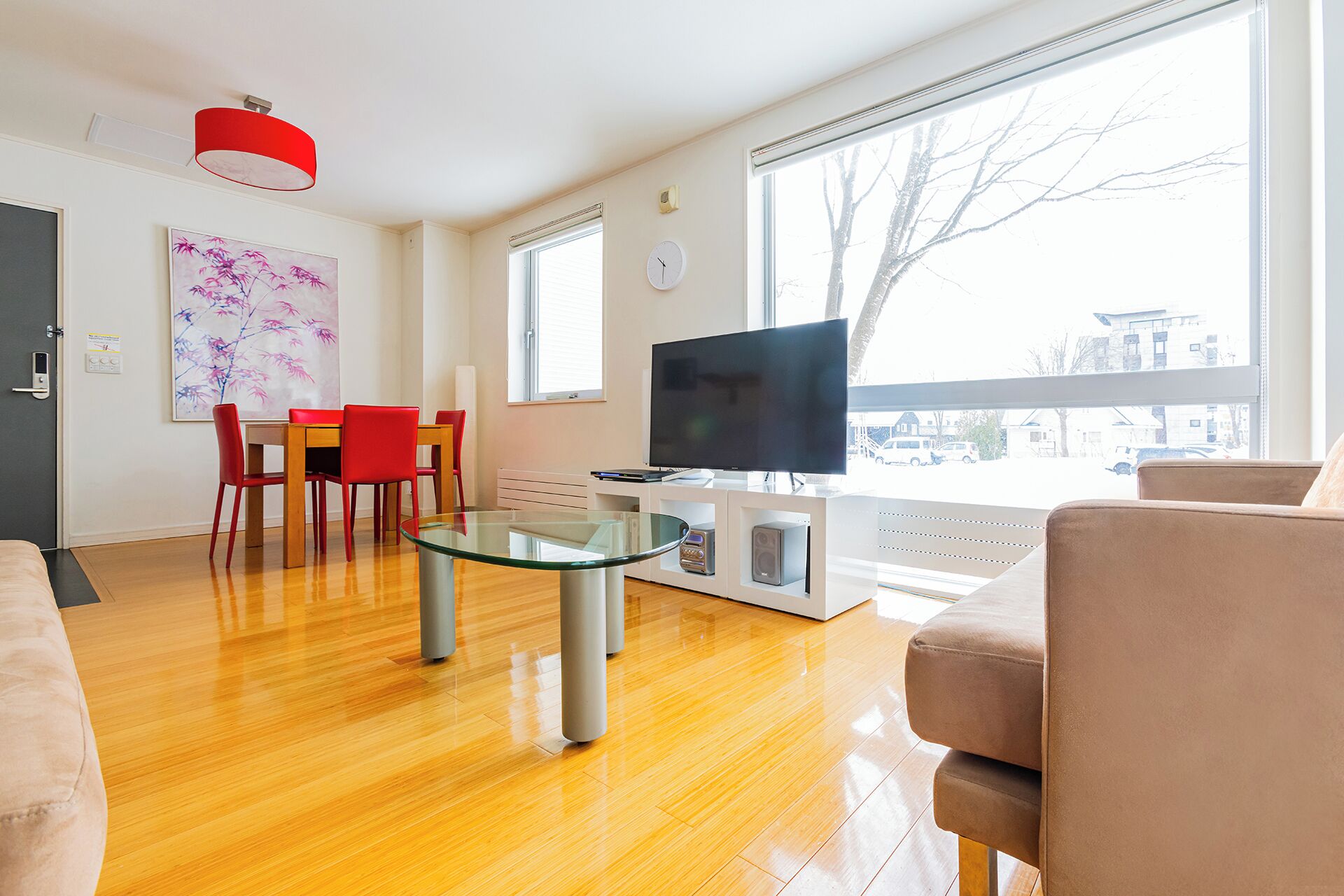 A modern living room with a wooden floor, a glass coffee table, a flat-screen TV, a red chair, a beige sofa, and a large window offering a view of the street.