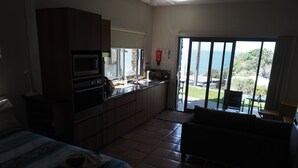 A compact kitchen with a microwave, stove, and sink, a dining area with a table and chairs, and a large sliding glass door leading to a balcony with a view of the sea. - WAVEBREAKER Scotts Head (Scotts Head)