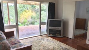 A living room with a sofa, a television, and a sliding glass door leading to an outdoor deck. - The Little Cottage 3 minutes walk to beach. Beds for 5. (Henderson Bay)