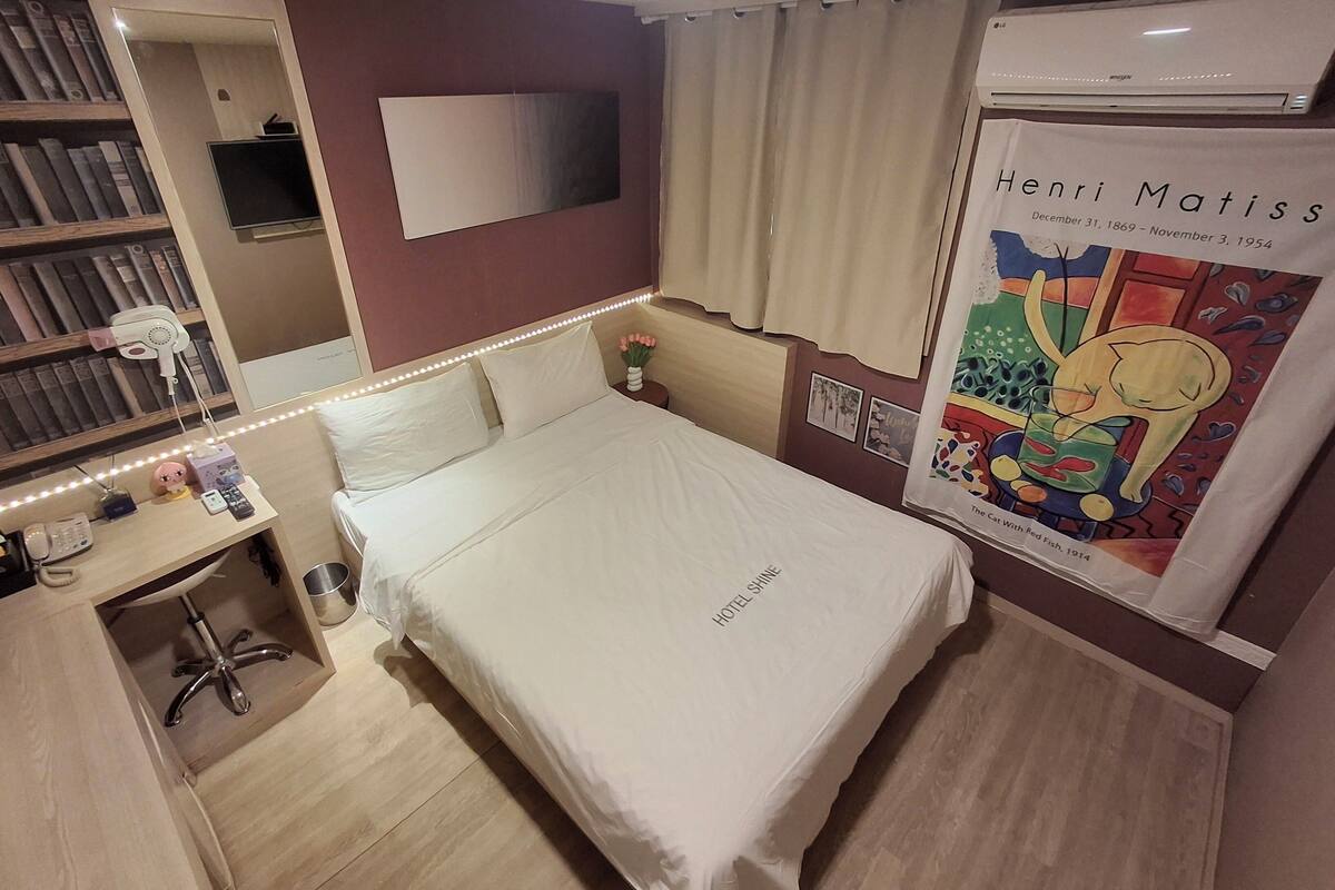 A hotel room with a neatly made bed, a bedside table with a lamp, a mirror, a bookshelf, and a poster of Henri Matisse's artwork.