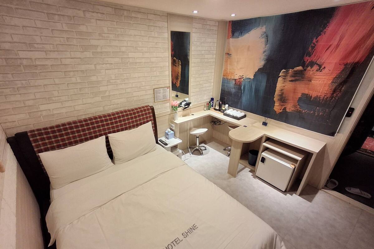 A hotel room with a bed, a desk, a mirror, and a large abstract painting on the wall.