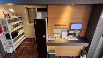 Un mostrador de recepción con un cartel que dice "SHINE STAY INFORMATION" y un monitor de computadora. Hay un cubo de basura y una planta cerca del mostrador.
