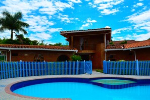 A swimming pool with a blue tiled border, surrounded by a blue fence, and a brick building with a red-tiled roof in the background. - Hostal Valle De Las Camelias (Pereira)