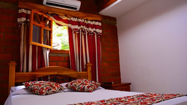 A bedroom with a wooden bed, patterned pillows, a window with red curtains, and a brick wall. - Hostal Valle De Las Camelias (Pereira)