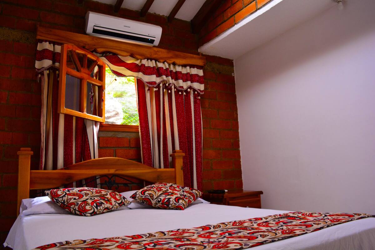 Ein Schlafzimmer mit einem hölzernen Bett, gemusterten Kissen, einem Fenster mit roten Vorhängen und einer Ziegelwand.