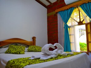 A bed with green patterned pillows, a white towel, and a wooden headboard in a room with a brick wall and a window with blue curtains. - Hostal Valle De Las Camelias (Pereira)