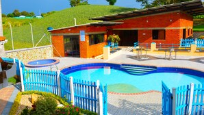 A swimming pool with a blue fence, a red building with a covered area, and a green hill in the background. - Hostal Valle De Las Camelias (Pereira)