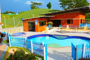 A swimming pool with a blue fence, a red building with a covered area, and a green hill in the background. - Hostal Valle De Las Camelias (Pereira)