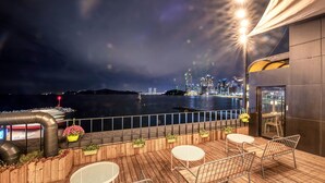 A wooden deck with outdoor seating, overlooking a body of water with city lights in the distance. - Mipo Oceanside Hotel (Busan)