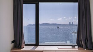 A room with a large window offering a view of a body of water and distant buildings. - Mipo Oceanside Hotel (Busan)