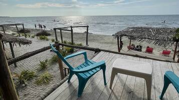 Terraza de madera con sillas y una mesa con vista a una playa y una estructura de techo de paja.