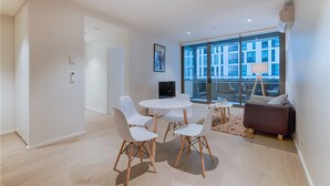 A modern living room with a white round table and chairs, a sofa, and a large window offering a view of buildings. - APC on Collins (Docklands)