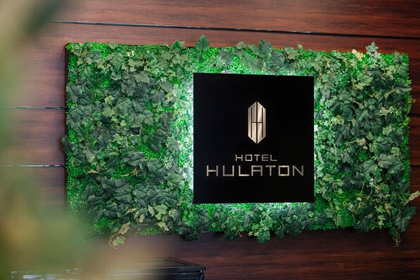 Un letrero enmarco con el texto 'HOTEL HULATON' y un logotipo geométrico, rodeado por un borde de hojas verdes.