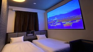 Ein Hotelzimmer mit zwei Betten, einem großen Fenster mit Stadtbild-Projektion, einer Deckenleuchte und einer Steckdose an der Wand.