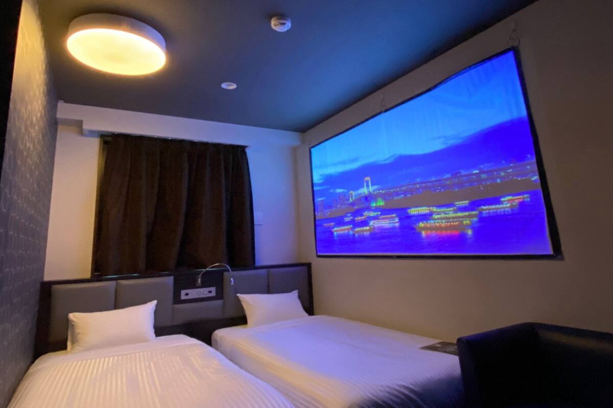 Ein Hotelzimmer mit zwei Betten, einem großen Fenster mit Stadtbild-Projektion, einer Deckenleuchte und einer Steckdose an der Wand.