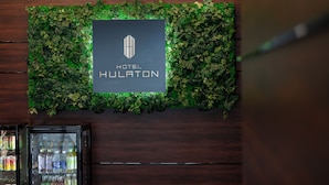 Ein an der Wand befestigtes Schild mit dem Schriftzug 'HOTEL HULATON' und einem Logo, umgeben von Grünpflanzen.