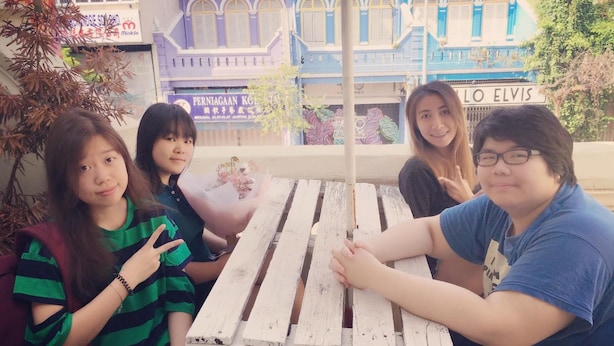 Four people sitting at a wooden table with a view of a building with arched windows and a sign that reads "LO ELVIS". - Vloft Backpackers Hostel (Ipoh)