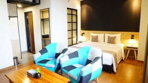 Quarto de hotel com uma cama, duas poltronas, uma mesa de madeira e uma estante fixada na parede.
