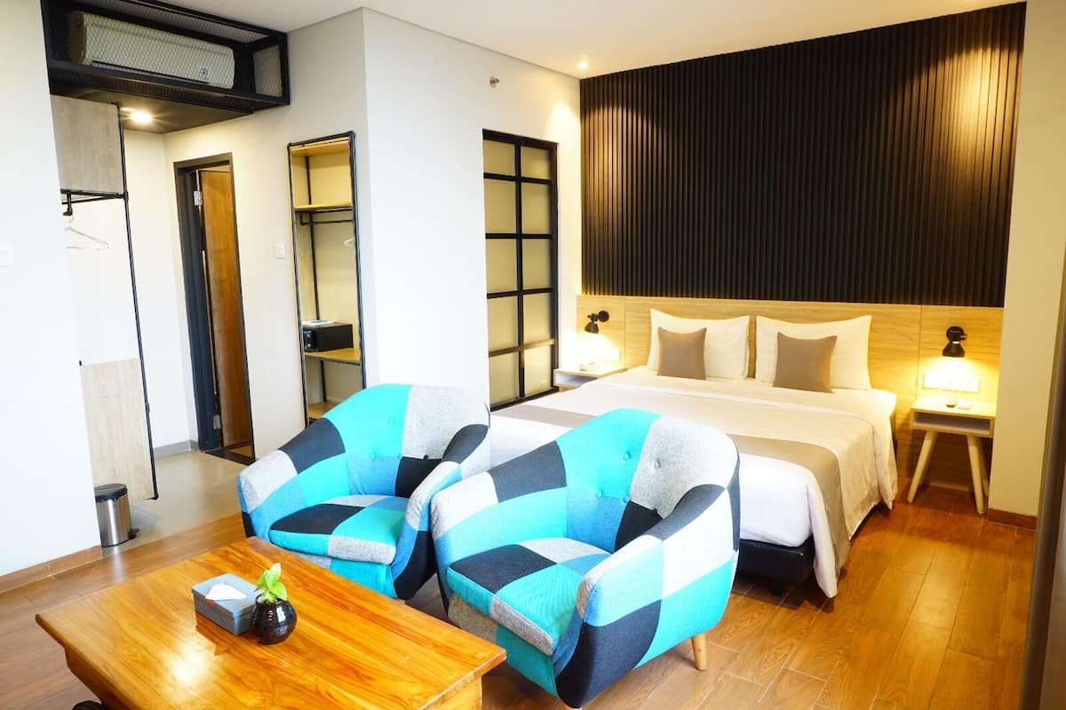 A hotel room with a bed, two armchairs, a wooden table, and a wall-mounted shelf.