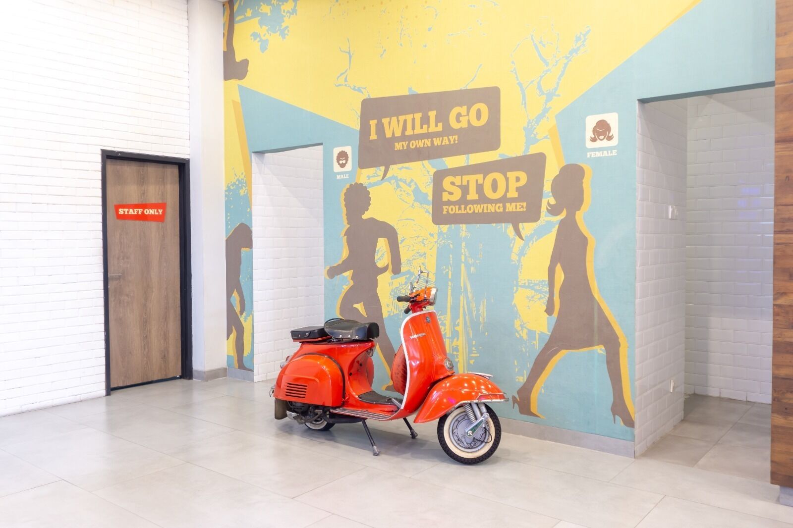 Uno scooter rosso parcheggiato in un corridoio con un murale raffigurante silhouette e frasi motivazionali.