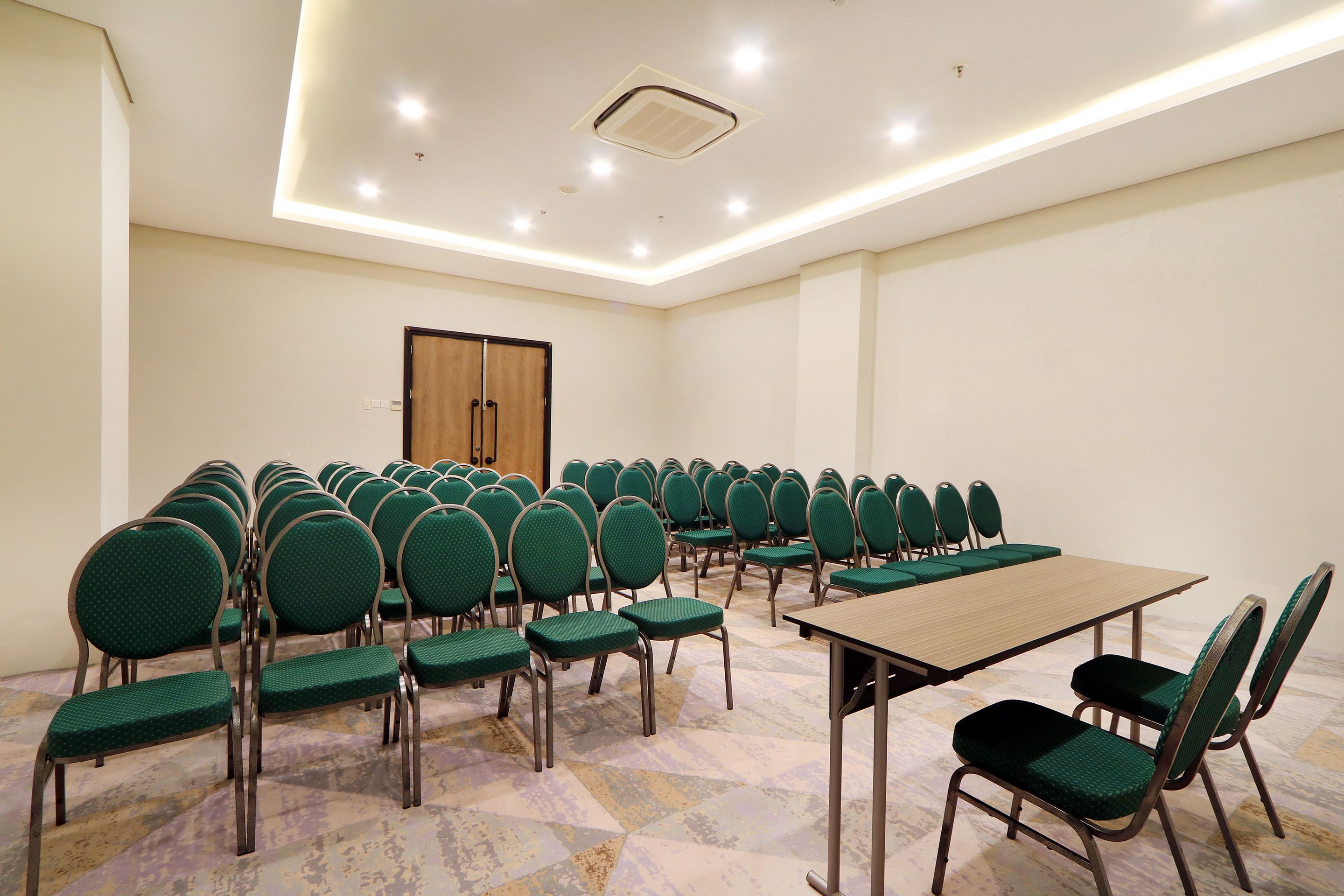 Une salle de conférence avec des rangées de chaises vertes, une longue table et une porte en bois.